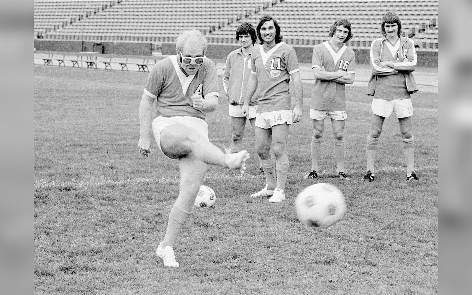 1976: Elton John avfyrar ett skott i egenskap av nybliven delägare av fotbollsklubben Los Angeles Aztecs. I bakgrunden skymtas stjärnförvärvet George Best. Elton John är en passionerad fotbollssupporter, är nära vän med Pelé och är gudfar åt David och Victoria Beckhams äldsta son Brooklyn. Foto: TT