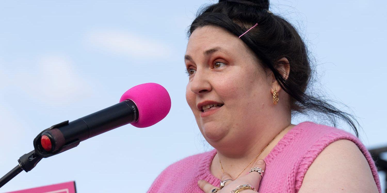 Lena Dunham står vid en rosa mik i rosa dress och handen på hjärtat. 