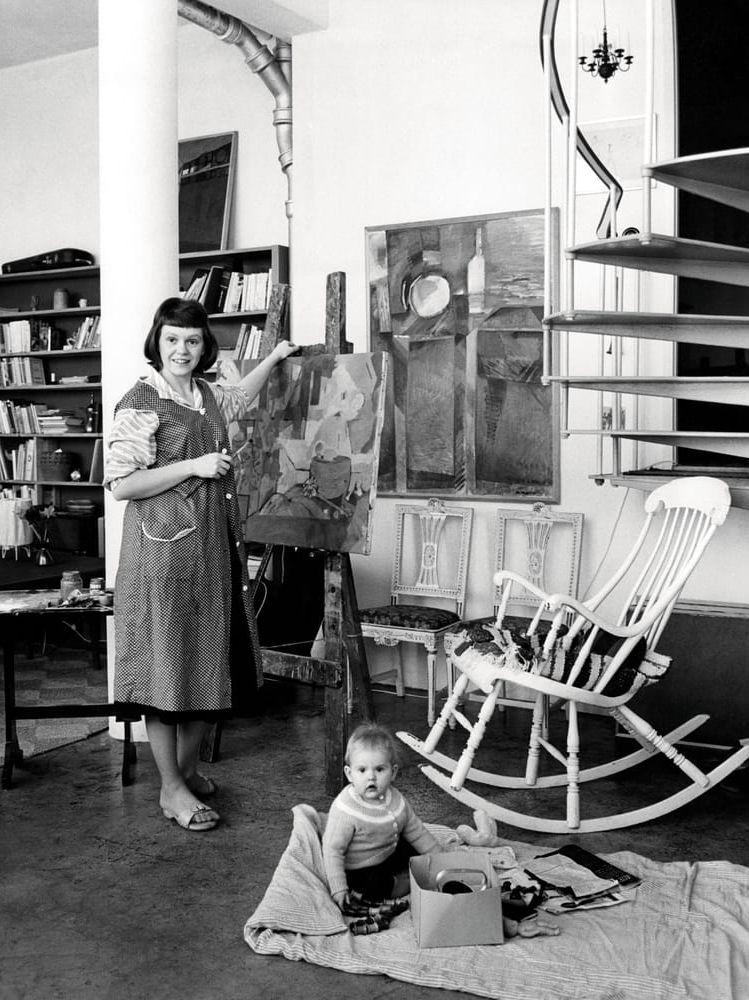 Konstnären Denice Zetterquist i ateljélägenheten i Biskopsgården 1960. Konstnärsparet Denice och Olle Zetterquist hörde till pionjärgenerationen i Biskopsgården. Denice Zetterquist (1929-2014) hade sedermera en mycket framgångsrik karriär som målare och grafiker, men tycks aldrig ha glömt sin kärlek till Göteborgs ytterstadsdelar – sålunda svarade hon för konstnärlig utsmyckning både i Tynneredsskolans aula och på O’Hareflygplatsen i Chicago. Ettåringen på golvet heter Nina Zetterquist. Även hon blev konstnär. Hon har ljusa minnen av åren i Biskopsgården:<em id="emphasis-8a9393a0ceb6f026dbb77ab858eb5a1f">Så många vuxna runt om kring mig som vi kände och så många kompisar. Ateljélägenheterna ligger ju på bottenvåningen i en lång rad. I varje lägenhet fanns det ett stort förråd och en lång gång band ihop förråden. Här brukade allas dörrar stå öppna när det var fester och det var det ofta, med mycket jazzmusik. Om någon av oss barn vaknade, visste vi att det bara var att gå ut i korridoren och höra från vilken öppen dörr det kom musik ...</em>