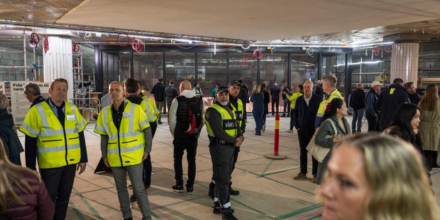 En samling människor i signalvästar visas runt i en nästan färdigbyggd underjordisk tågstation.