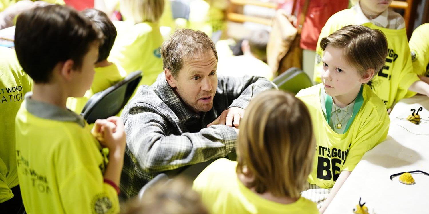 Sebastian Vettel sitter på huk iklädd grårutig skjorta omgivna av lågstadiebarn i ett klassrum.