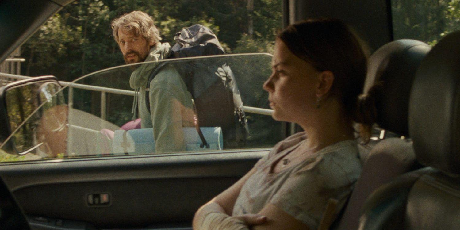 Hedvig Nilsson i rollen som dottern i filmen sitter i en bil och tittar surt framför sig medan Adam Lundgren i rollen som fadern står utanför och sneglar oroligt på henne. 