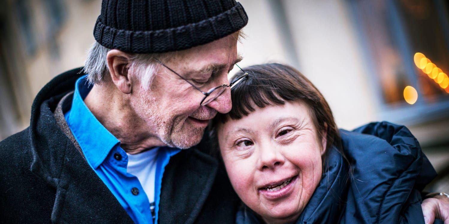 Man i rock, skjorta, glasögon och mössa håller om kvinna i blå jacka.