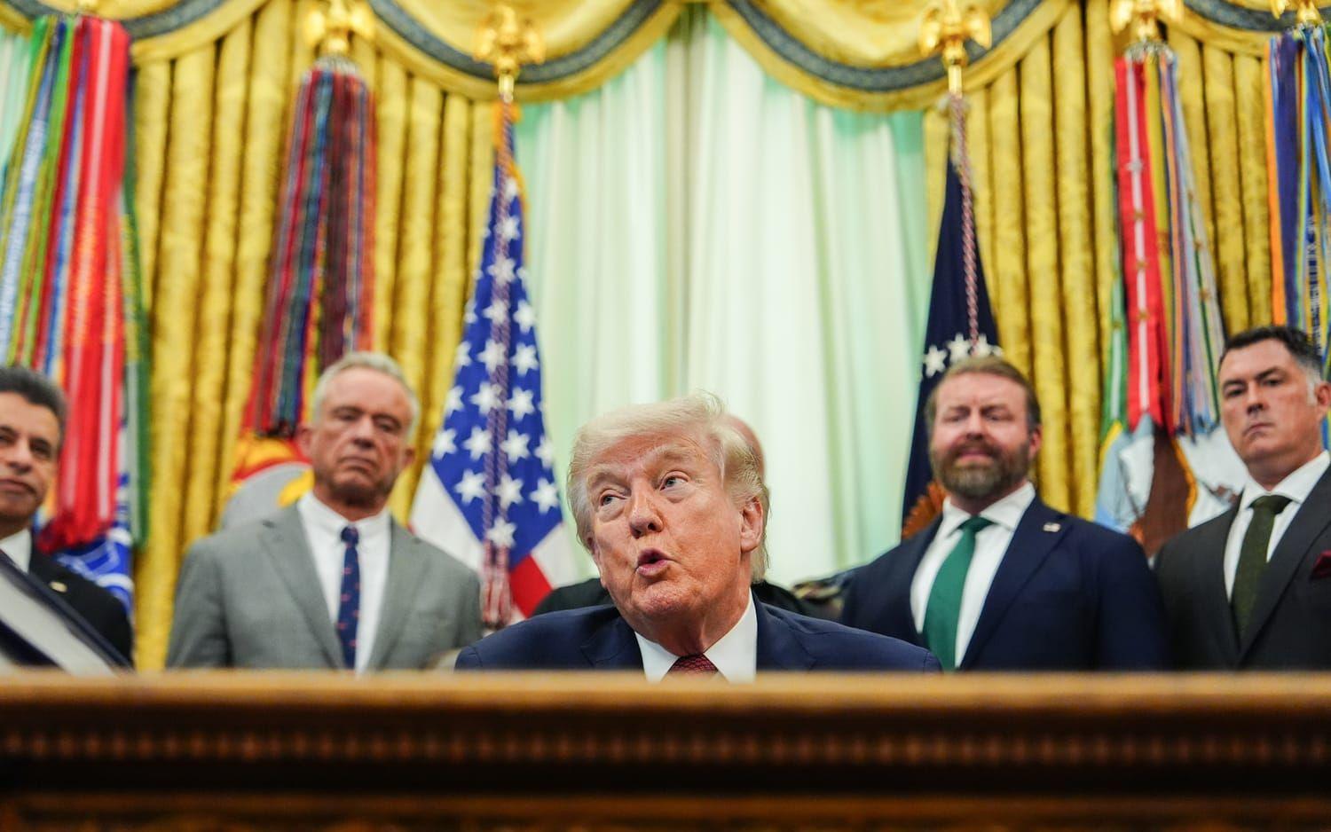 Man med blont hår sitter vid skrivbord, i bakgrunden flera män som står upp.