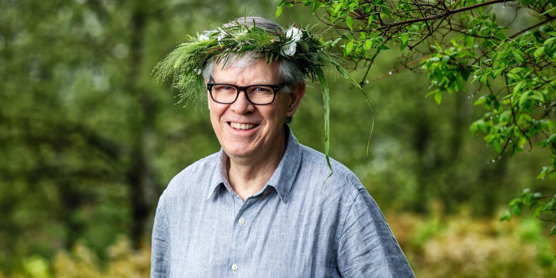 Biologen och naturjournalisten Henrik Ekman poserar inför Sommar i P1. Han har på sig den traditionsenliga kransen och en blå skjorta. Han bär glasögon och ler brett.