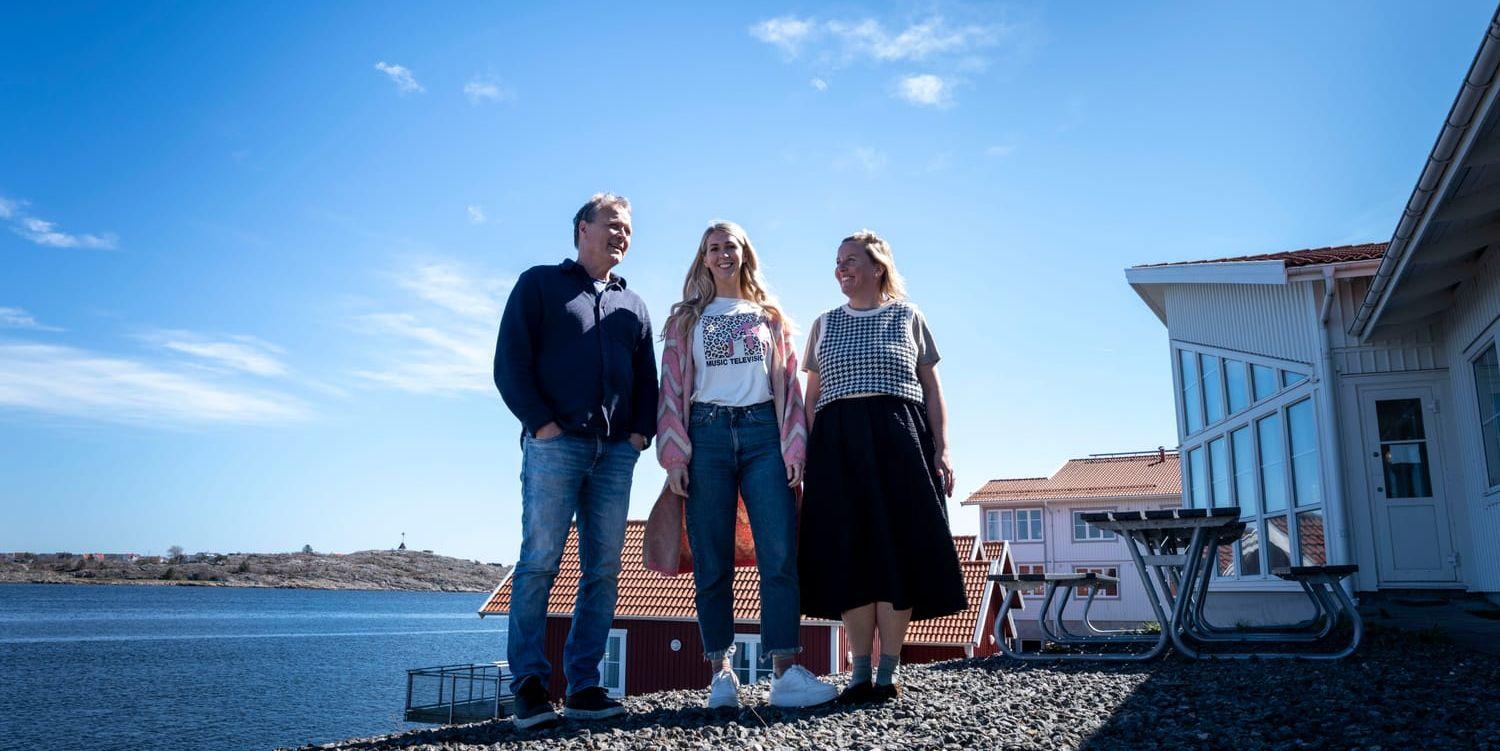 Tre personer står på grusplan. Vit skolbyggnad, hav och röda fiskebodar i bakgrunden.