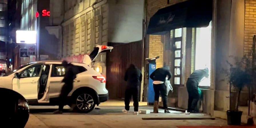 En bil med bakluckan öppen, flera svartklädda personer som står vid ett sönderslaget skyltfönster.