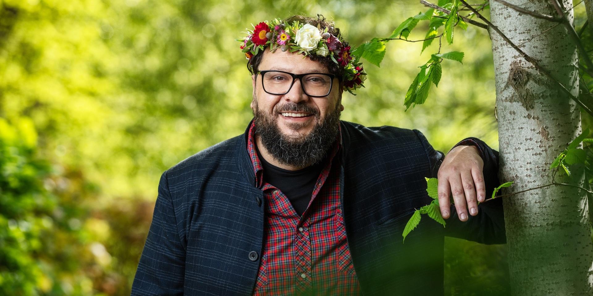 Filmregissören Goran Kapetanović poserar för Sommar i P1. Han lutar sig mot en björk. Han har blomsterkrans. Randig mörkblå overshirt över en rödrutig skjorta och svart t-shirt. Han har ett rejält skägg och stora glasögon med svarta bågar.