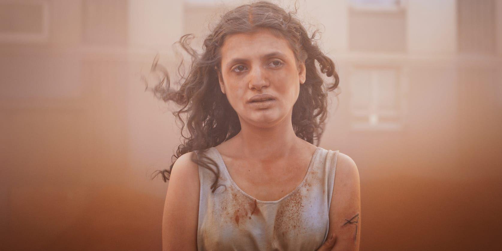 En 13-årig flicka (Alpha) i smutsigt vitt linne och långt mörkt hår står utanför ett hyreshus i en röd vind av sand och grus som lagt sig i en hinna över hennes ansikte.