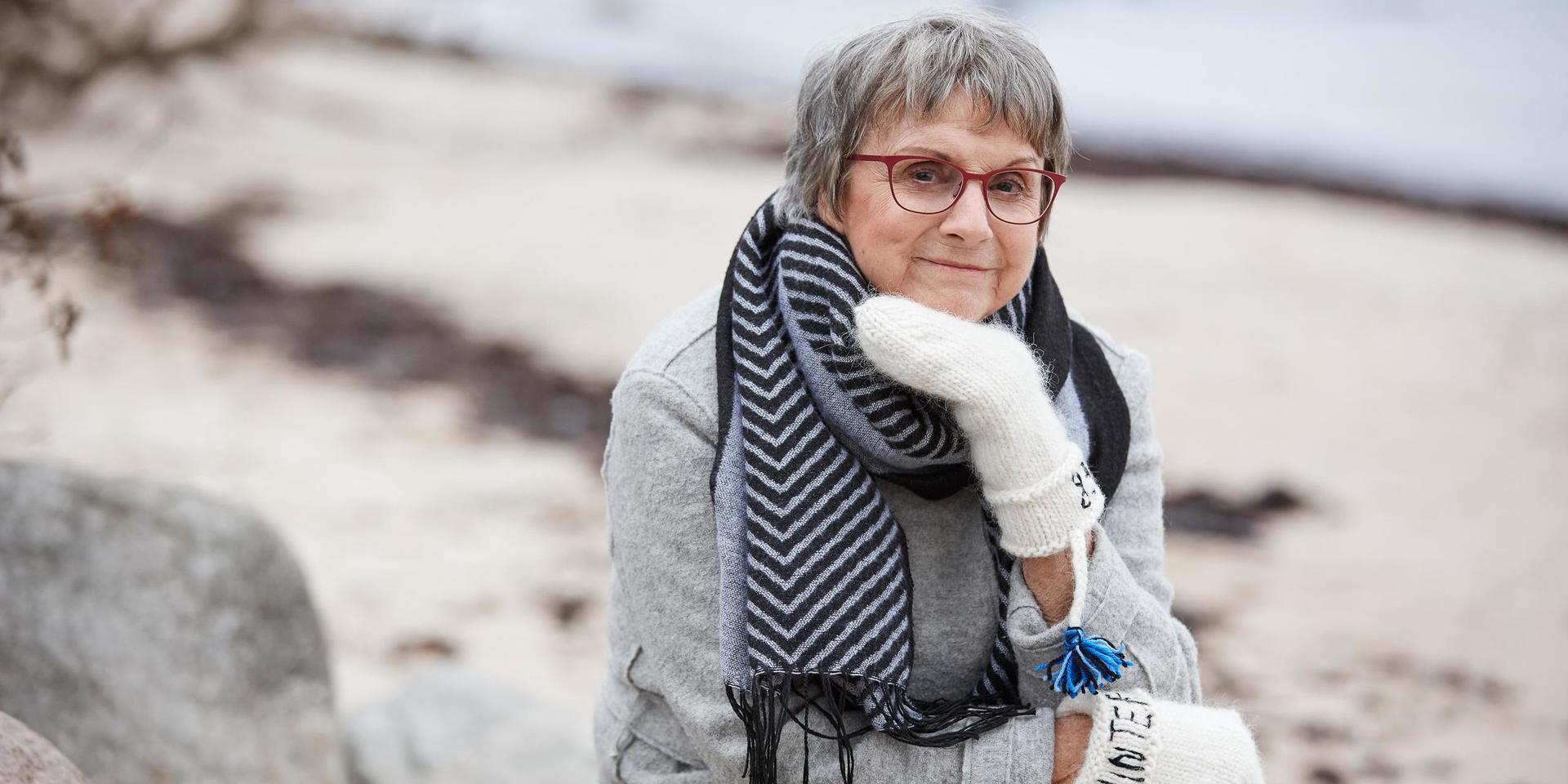 Psykologen och författaren Patricia Tudor Sandahl sitter med röda glasögon, vita vantar och en randig halsduk. Hon sitter på ett gäng klippblock vid stranden för en fotografering för Sveriges Radios program Vinter i P1.