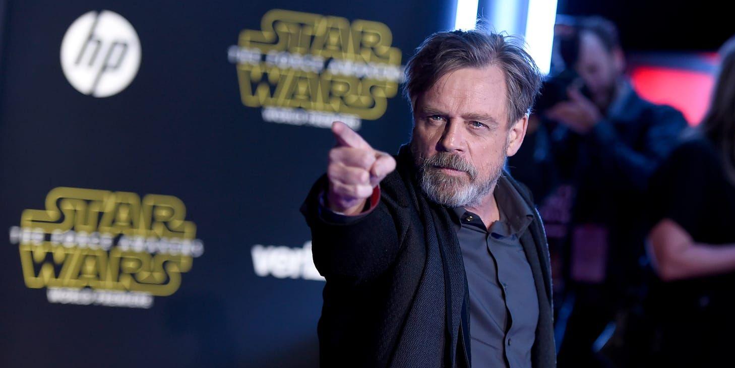 Skådespelaren Mark Hamill i skägg med svart skjorta och svart kavaj på röda mattan där han kisar med ögonen och pekar på något med hela armen.