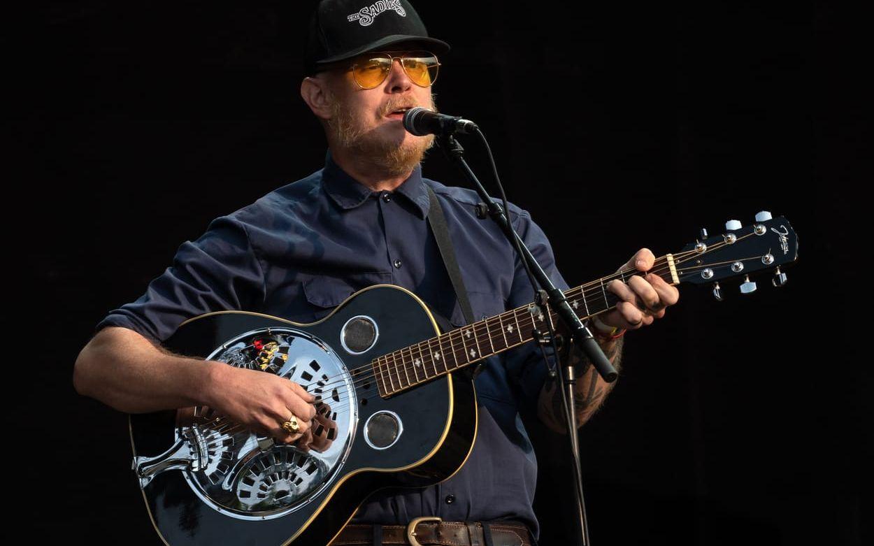 Taylor Kirk vid en mikrofon på scen, med akustisk gitarr.