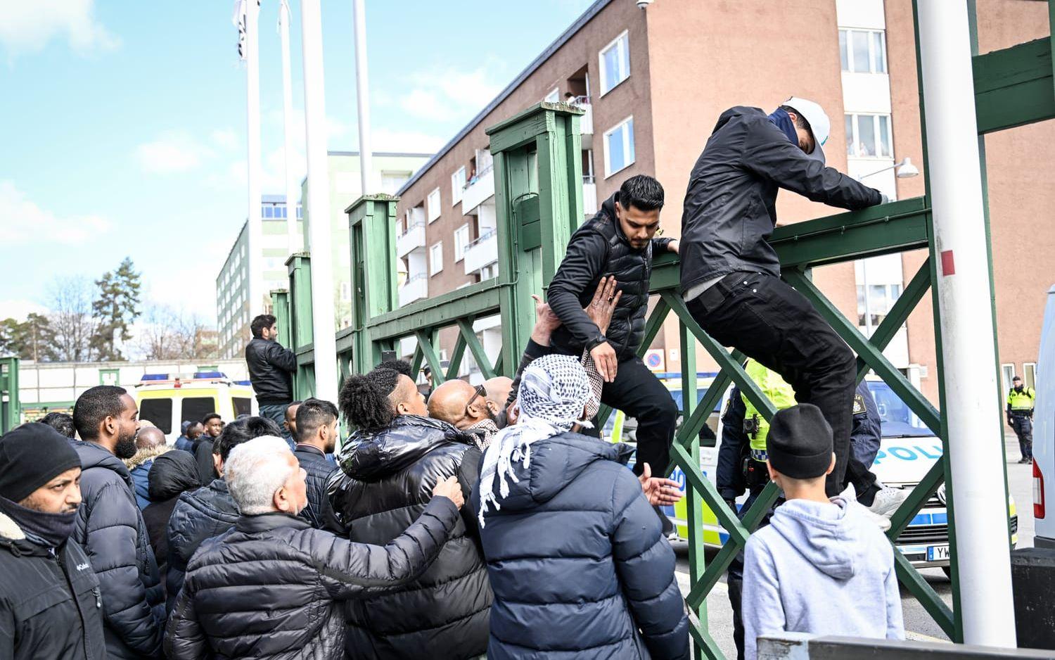 Människor försöker lugna upprörda män och få dem att klättra ner från ett stängsel i Stockholmsförorten Rinkeby där Rasmus Paludan manifesterar med koranbränning på långfredagen.