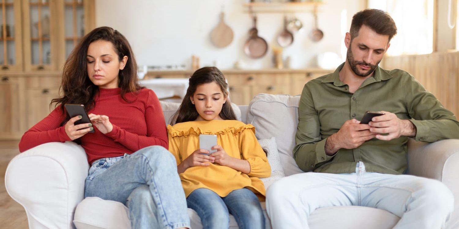 Familj med mamma pappa och dotter sitter i en soffa och stirrar på sina telefoner. 